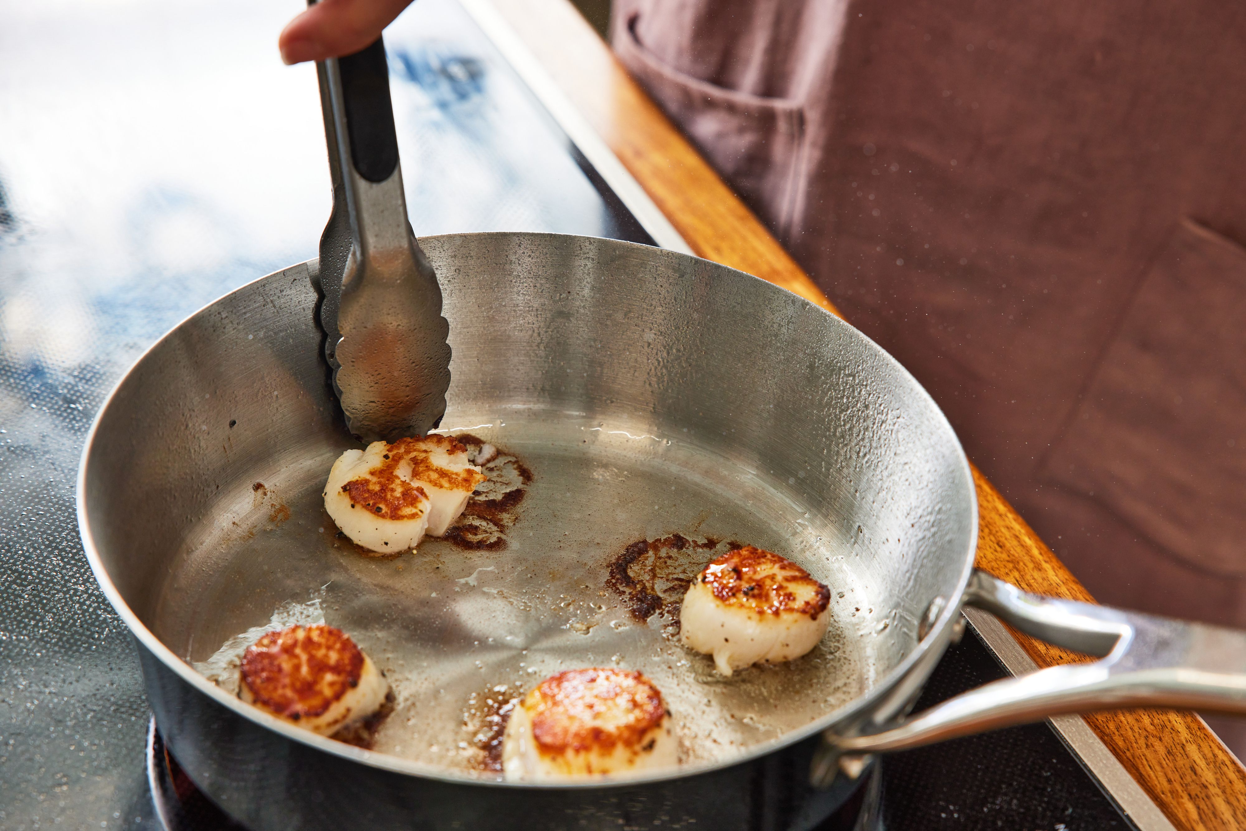 Person's hand using tongs while cooking scallops on a pan from the Anolon Nouvelle Stainless 10-Piece Cookware Set