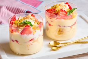 Two glasses of layered dessert with fruit and cream on a white tray with gold spoons