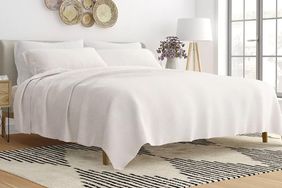 A neatly made bed with white sheets and a decorative rug underneath featuring wallmounted plates and a lamp in the background