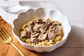 A dish of beef stroganoff served in a white bowl on a table with a gold fork and an orange napkin
